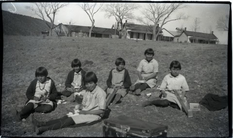 Six girls sit on a grassy field eating lunch, with leafless trees and several buildings in the background. The scene appears to be in a rural setting.